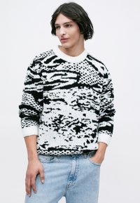 Suéter de punto con un patrón en blanco y negro, con cuello redondo y puños de canalé, llevado sobre unos jeans de un azul claro. Textura suave y corte relajado.