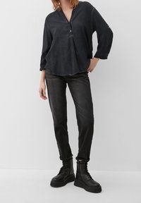 Donna che indossa una blusa nera a maniche lunghe, jeans grigio scuro e stivali platform neri con lacci, in piedi contro uno sfondo bianco.