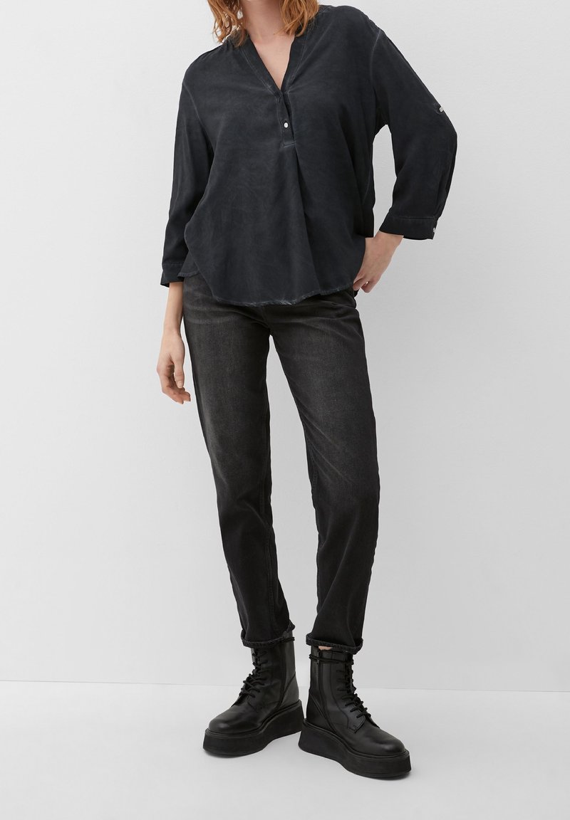 Donna che indossa una blusa nera a maniche lunghe, jeans grigio scuro e stivali platform neri con lacci, in piedi contro uno sfondo bianco.