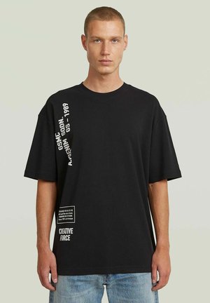Młody mężczyzna z krótkimi włosami, noszący czarny oversize T-shirt z białym pionowym i dolnym tekstem, stojący na jednolitym jasnym tle.