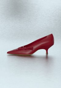 Chaussure rouge en cuir à talon kitten, bout pointu et nœud décoratif sur le dessus, sur fond gris clair.
