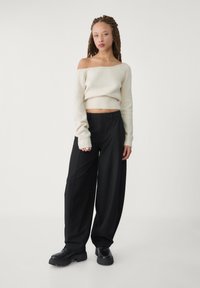 Pull en maille crème à épaules dénudées, ourlet côtelé, associé à un pantalon noir ample et des bottes noires épaisses, sur un fond neutre.