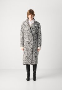 Abrigo largo en blanco y negro de patrón de espiga, con un amplio cuello, diseño de doble botonadura y puños en crema que contrastan, presentando un ajuste relajado.