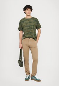 T-shirt verde mimetico a maniche corte, pantaloni slim-fit beige, sneakers verdi e bianchi, e una borsa a tracolla verde. Sfondo semplice.