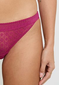 Perizoma fucsia in pizzo con fascia in vita ondulata, caratterizzato da motivi floreali intricati e un design trasparente e testurizzato sul tessuto.