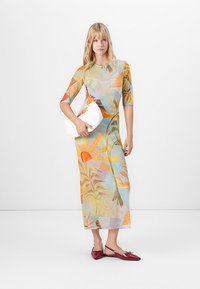 Vrouw in een kleurrijke bloemenmaxi-jurk, met een witte clutch in de hand en rode puntige flats, staande tegen een effen witte achtergrond.