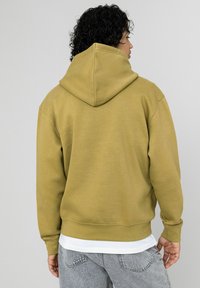 Sweat à capuche vert olive avec cordon de serrage, poignets et ourlet côtelés. Porté par-dessus une chemise blanche, associé à un jean gris. La texture semble douce.