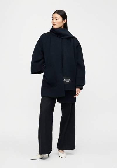 Γυναίκα που φοράει υπερμεγέθη σκούρο μπλε παλτό με λεπτομέρεια φουλαριού, φαρδιά μαύρη παντελόνα και λευκά μυτερά τακούνια, μπροστά σε απλό φωτεινό φόντο.