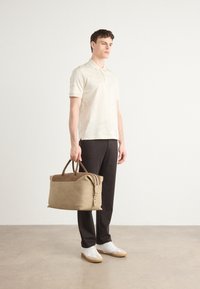 Smėlio spalvos sportinė kuprinė su rudos odos akcentais, turinti dvi rankenas ir užtrauktuką. Modelis vilki šviesią polo marškinėlį ir tamsias kelnes, avėdami baltus batus.