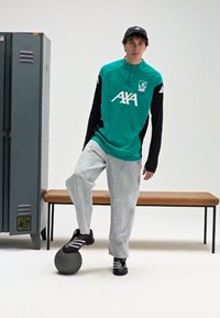 Turkio ir juodos spalvų sportinis užsegamas megztinis su logotipu, šviesios džinsinės kelnės, juodi sportbačiai ir pilkas kamuolys, pozuojama prie spintelės ir suolo.