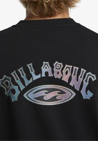 Sudadera negra con un gráfico brillante e iridiscente de "Billabong" y un logo en el centro. Presenta cuello acanalado y tejido texturizado.