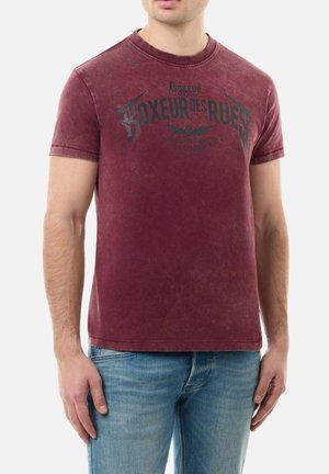 Man wearing faded burgundy short-sleeve t-shirt with "Boxeur Des Rues" text and logo, paired with light blue jeans against white background.