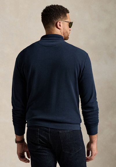 Μπλε ναυτικό μακρυμάνικο πόλο με γιακά όρθιο, ριμπ μανσέτες και απαλή υφή, σε συνδυασμό με σκουρόχρωμο τζιν. Τα γυαλιά ηλίου προσθέτουν μια μοντέρνα λεπτομέρεια.