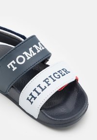 Navyja i biele šľapky so zriediteľnými popruhmi, na ktorých sú nápisy "TOMMY" a "HILFIGER", vyrobené z syntetického materiálu s textúrovanou podrážkou.