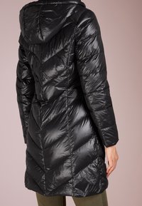 Svart quiltad pufferjacka med huva och chevronstickning, bärs av en person som står mot en enfärgad bakgrund.