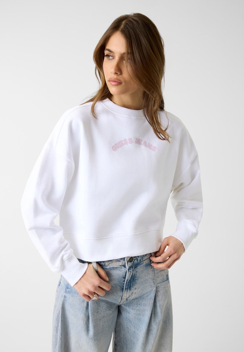 Jeune femme aux cheveux châtain clair portant un sweat-shirt blanc avec « Guess Jeans » en rose et un jean bleu délavé, regardant vers sa gauche.