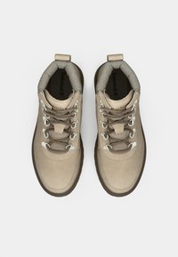 Beige tursko med mokka- og tekstiloverdel, rund tå, polstret krave og grå snører. Metalløyer og en mørkebrun gummisåle.