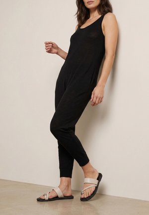 Femme portant une combinaison noire sans manches et des sandales beige à enfiler avec des bagues d'orteils, debout contre un mur et un sol beige clair.