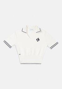 Haut en maille blanc à manches courtes avec bordure noire sur le col et les poignets, un petit patch logo noir et blanc sur la poitrine, et un emblème crocodile vert près de l'ourlet.