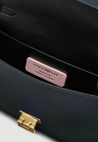 Interno di una borsa in pelle nera con un'etichetta rosa chiaro su cui è scritto "COCCINELLE - Pelle Italiana Autentica," con un dettaglio della chiusura dorata.