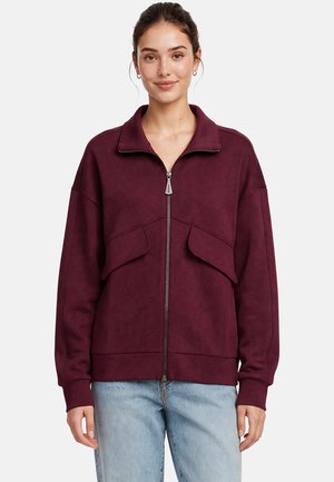 Donna che indossa una giacca bordeaux con cerniera e tasche con patta e jeans azzurri chiari, in piedi e rivolta frontalmente su uno sfondo semplice.