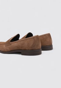 Mocassins en daim marron avec un bout arrondi, un design à enfiler et une semelle en caoutchouc basse. Présente des coutures visibles et une texture lisse.