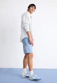 Camisa a rayas blanca y azul claro, pantalones cortos de mezclilla claros, zapatillas azul y blancas con acentos grises, de pie sobre un suelo azul.