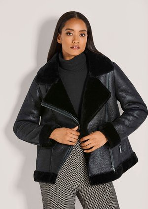 Frau trägt eine schwarze Lammfelljacke über einem schwarzen Rollkragenpullover und gemusterten Hosen, sie stellt den vorderen Reißverschluss der Jacke ein.