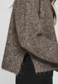 Brūna adīta džemperis ar teksturētu, pūkainu izskatu. Tam ir brīvs piegriezums un sānu šķirbes, kas nodrošina ērtu kustību.