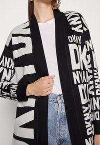 Svart och krämfärgad stickad cardigan med tydlig "DKNY" text, öppen framsida och ribbade detaljer. Bärs över en vit topp och ljus denimjeans.