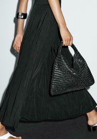Woven black bag with a triangular shape, textured surface, and sturdy handle, paired with a flowing black skirt and white sandals.