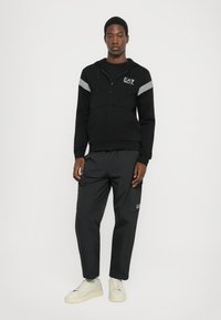 Juoda zip-up džemperis su baltomis juostelėmis pečių srityje, derinamas su juodais cargo tipo kelnais. Ant krūtinės yra logotipas, o prie drabužių priderinti kreminiai sportbačiai.