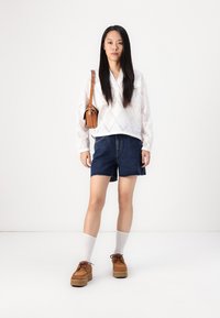 Vit långärmad skjorta med diamantformade utskärningar, mörka denimshorts, bruna platformskor och krämfärgade ankelsockor. Liten brun crossbody-väska.