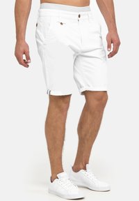 Man in witte knielange shorts met knoop- en ritssluiting en witte veterschoenen op een effen achtergrond.