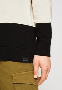 Jersey de punto en rayas crema y negro, con puños acanalados y una etiqueta de marca en el dobladillo. Mano cerca de los pantalones cargo color caqui.