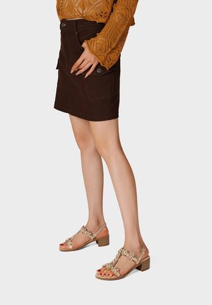 Donna che indossa una minigonna marrone con tasche frontali, un top in pizzo color senape a maniche lunghe e sandali beige con tacco e decorazioni floreali.