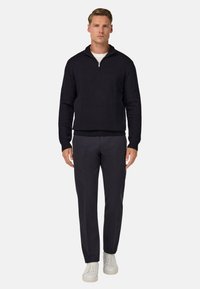 Jūras spēku krāsas teksturēts puloveris ar ceturtdaļas rāvējslēdzēju, uzvilkts virs baltas krekla. Apvienots ar pielāgotām tumšām biksēm un baltām sporta apavām.