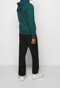 Türkise Kapuzenjacke mit Kordelzug, kombiniert mit schwarzen Hosen und weißen Turnschuhen. Das Outfit hat eine lockere Passform und eine glatte Textur.