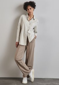 Camicia a righe crema e oliva, abbinata a pantaloni beige a gamba larga e sneakers bianche. Modello in piedi contro un muro grigio.