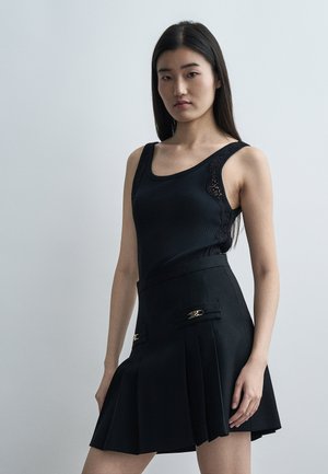 Ung kvinde med langt mørkt hår iført en sort ærmeløs top med blonder og et plisseret sort nederdel med gulddetaljer.