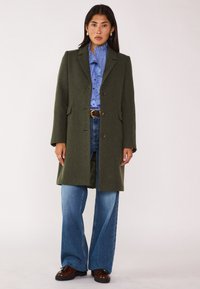 Cappotto di lana verde oliva con vestibilità sartoriale, design monopetto, rever a punta e due tasche frontali. Abbinato a una camicia a righe blu e jeans.