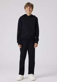 Zwarte sweatshirt met capuchon en bijpassende zwarte broek, beide van een zachte stof, en witte sneakers, staande tegen een lichte achtergrond.