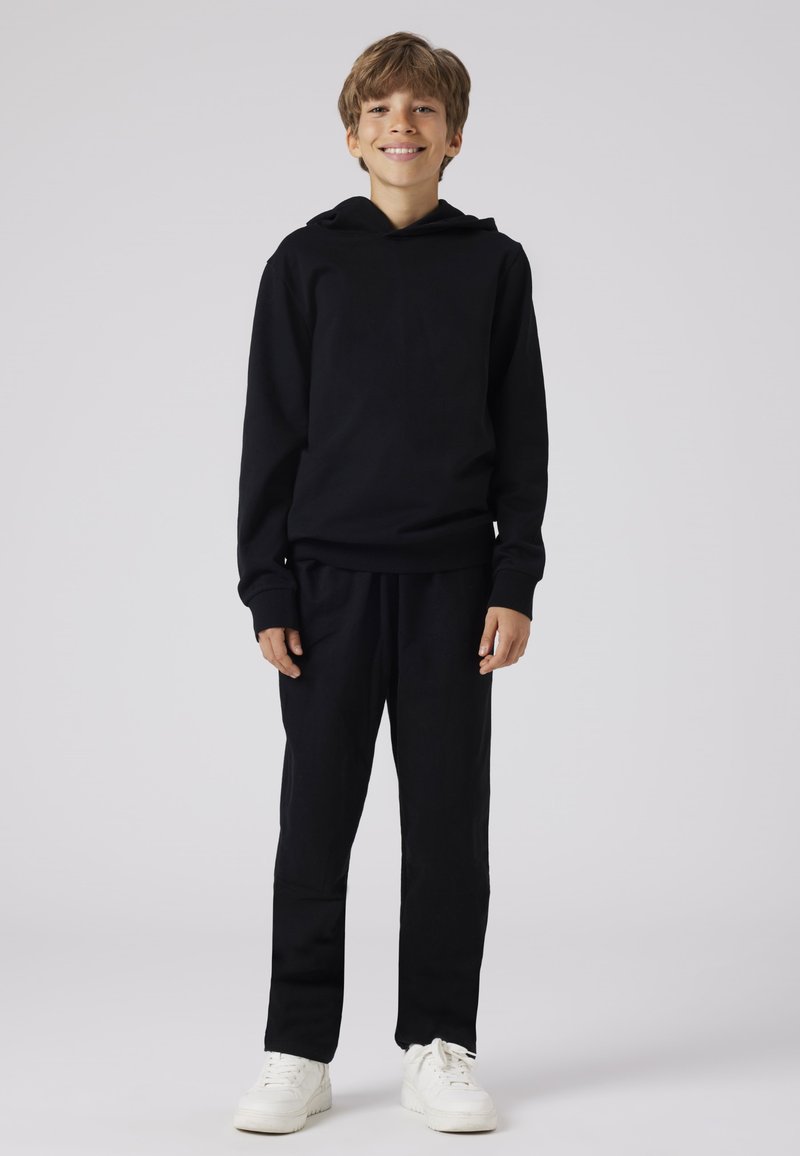 Zwarte sweatshirt met capuchon en bijpassende zwarte broek, beide van een zachte stof, en witte sneakers, staande tegen een lichte achtergrond.