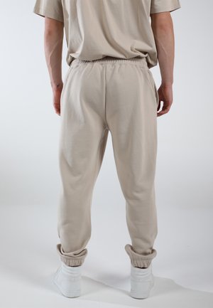 Beige Baumwoll-Jogginghosen mit elastischem Bund und Bündchen, kombiniert mit weißen Sportschuhen. Der Stoff hat eine weiche Textur und eine lockere Passform.
