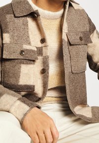 Mand iført beige og brunternet uldjakke over en creme- og gulstrikket sweater med cremefarvede bukser, sidder med hånden hvilende på knæet.