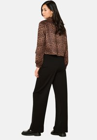 Zrezaná hnedá bunda s leopardím vzorom a dlhou rukávou, skombinovaná so širokými čiernymi nohavicami a čiernou obuvou, z pohľadu zozadu.