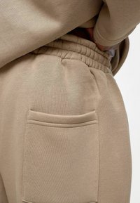 Beige katoenen joggingsbroek met een gathers elastische tailleband en een zijzak met een gestikte rand. Zachte textuur en een relaxed fit.