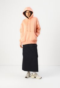 Sweat-shirt à capuche rose avec une poche frontale, associé à une longue jupe noire avec des poches cargo sur les côtés, et des chaussures de sport dans des tons neutres.