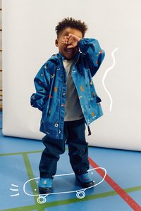Veste imperméable bleue avec des motifs colorés, associée à un pantalon bleu foncé et des chaussures bleues assorties, se tenant sur un graphique de skateboard.