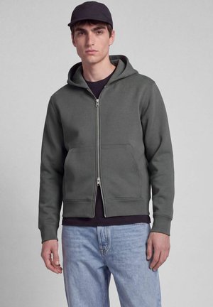 Jonge man met een donkere zip-up hoodie, zwarte pet, zwart shirt en lichtblauwe spijkerbroek, staand tegen een effen grijze achtergrond.